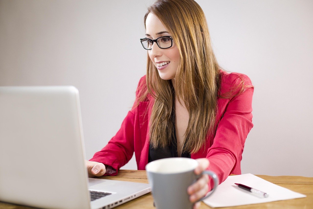A professional woman wearing glasses and a red blazer smiling while working on her laptop at a desk with a cup of coffee, representing a modern and focused business owner.